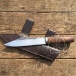 Carbon steel seax knife with a round wood handle lying across its dark brown leather sheath on weathered wooden boards