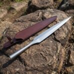 Forged steel spear head and burgundy leather sheath laid side by side on a large rock in warm sunlight