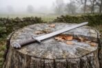 Hand forged steel Viking sword with leather wrapped grip resting across a tree stump with autumn leaves, misty field behind