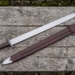 Hand forged steel Viking sword with leather wrapped grip laid on a wooden table beside a brown leather scabbard