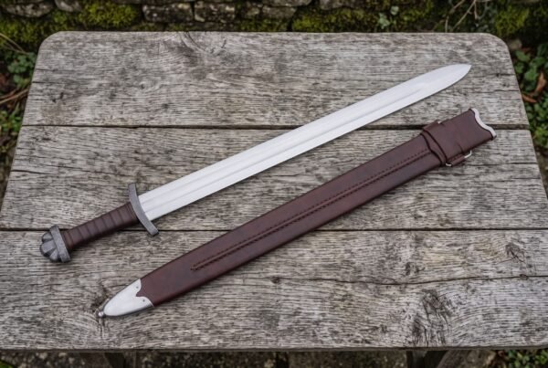 Hand forged steel Viking sword with leather wrapped grip laid on a wooden table beside a brown leather scabbard