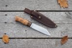 Plain steel seax knife with a wood handle laid on grey planks below its stitched brown leather sheath and autumn leaves