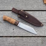 Plain steel seax knife with a wood handle laid on grey planks below its stitched brown leather sheath and autumn leaves