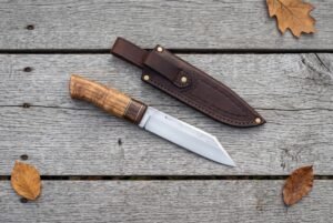 Plain steel seax knife with a wood handle laid on grey planks below its stitched brown leather sheath and autumn leaves