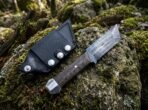 Satin steel tanto knife with dark wood handle and steel pommel cap lying beside its black leather sheath on a mossy rock
