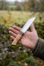 Seax knife in plain carbon steel with a figured wood handle resting on an open palm against a mossy stone wall
