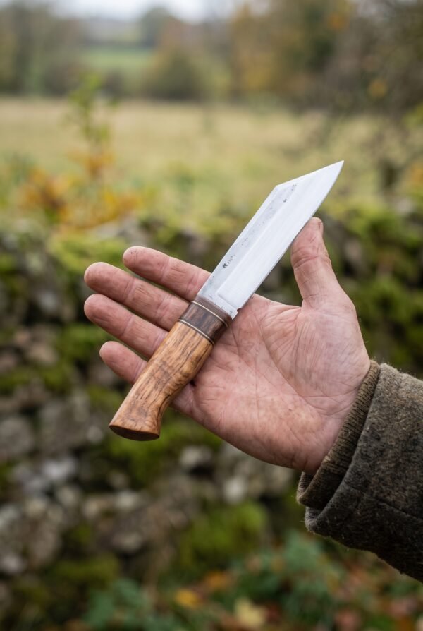 Seax knife in plain carbon steel with a figured wood handle resting on an open palm against a mossy stone wall
