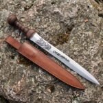 Short stainless steel sword with knotwork dragon etching and turned wood grip on a lichen covered rock beside a brown leather scabbard