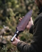 Steel spear head being slid into a dark brown leather sheath with a brass stud, held in both hands outdoors