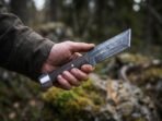 Steel tanto knife held flat in an open hand, side view of the angled point and wood handle over mossy boulders