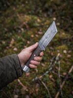 Steel tanto knife with an etched wave band on the blade held upright in one hand above a leafy forest floor