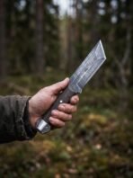 Steel tanto knife with dark wood grip and engraved pommel cap held up against a blurred pine forest