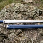 Stainless steel replica longsword with blue leather grip and curved guard lying on a lichen covered rock beside its blue scabbard