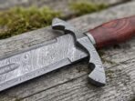 Close up of the Damascus guard and rosewood grip of a Viking sword resting on weathered grey wood with moss
