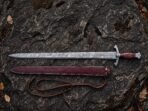 Damascus steel Viking sword with rosewood grip above its burgundy tooled leather scabbard and belt strap on dark rock