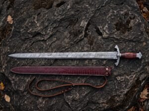 Damascus steel Viking sword with rosewood grip above its burgundy tooled leather scabbard and belt strap on dark rock