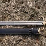 Damascus steel sword with brass cross guard and wrapped wood grip laid on stone above its black leather scabbard with brass fittings