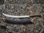 Stainless steel curved elven sword with brass guard and spiral wrapped grip beside its black scabbard on lichen covered rock