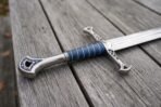 Stainless steel fantasy longsword hilt with blue wrapped grip and pierced silver guard resting on weathered grey boards