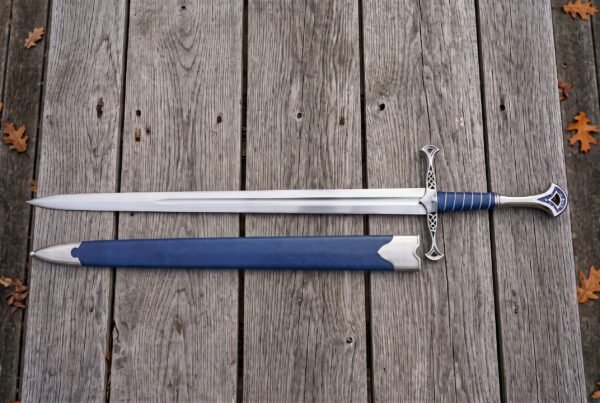 Stainless steel fantasy longsword laid beside a blue scabbard with silver chape on weathered boards with autumn leaves