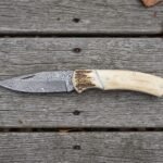 Damascus folding knife with camel bone handle and stag bolster lying open on grey wooden boards, full side view