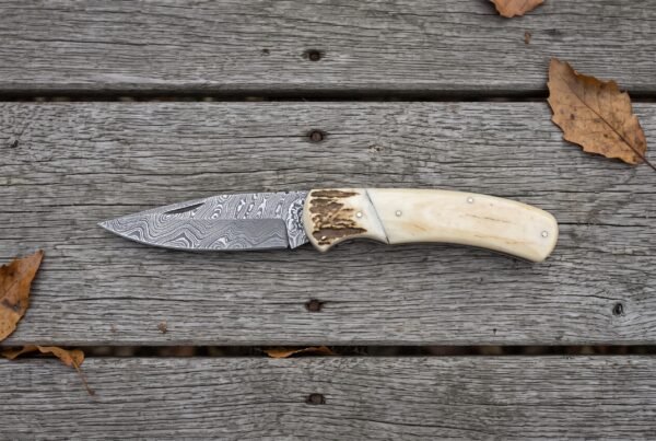Damascus folding knife with camel bone handle and stag bolster lying open on grey wooden boards, full side view