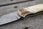 Close view of a Damascus folding knife with camel bone handle and stag bolster resting on weathered wood