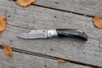 Damascus folding pocket knife with buffalo horn handle lying open on weathered wooden planks with scattered autumn leaves