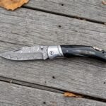 Damascus folding pocket knife with buffalo horn handle lying open on weathered wooden planks with scattered autumn leaves