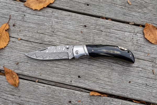 Damascus folding pocket knife with buffalo horn handle lying open on weathered wooden planks with scattered autumn leaves