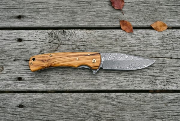 Damascus folding pocket knife with olive wood handle lying open on grey boards with autumn leaves, seen from above