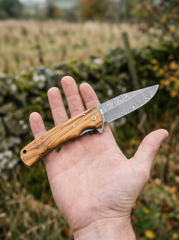 Damascus folding pocket knife with olive wood handle held open in a palm in front of a hedge and field