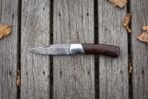 Damascus folding pocket knife with pakka wood handle and steel bolster lying open on grey planks, seen from above