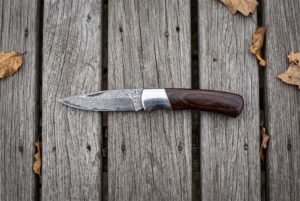 Damascus folding pocket knife with pakka wood handle and steel bolster lying open on grey planks, seen from above