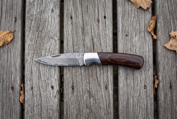 Damascus folding pocket knife with pakka wood handle and steel bolster lying open on grey planks, seen from above