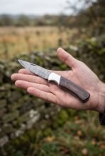 Damascus folding pocket knife with pakka wood handle and steel bolster held open in a hand outdoors near a stone wall