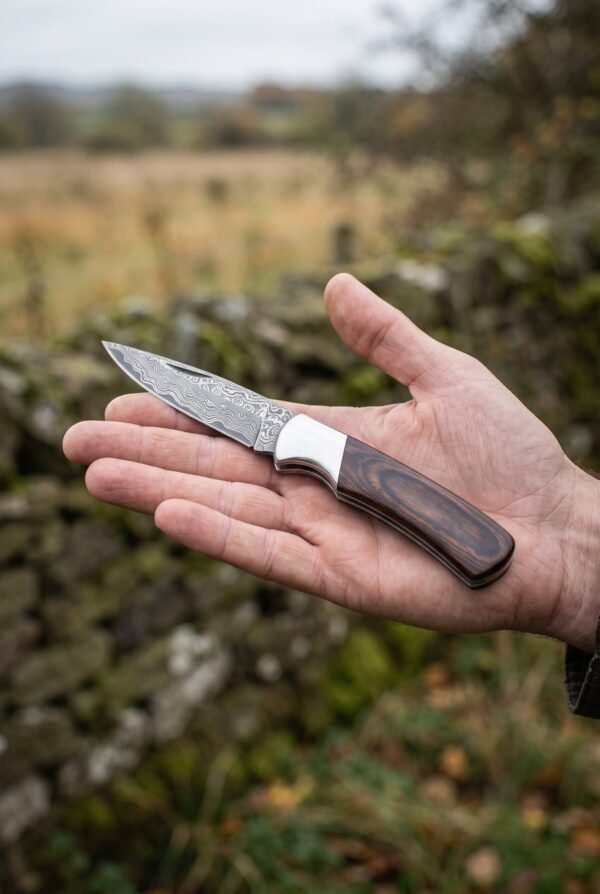 Damascus folding pocket knife with pakka wood handle and steel bolster held open in a hand outdoors near a stone wall