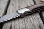 Close view of a Damascus folding pocket knife with steel bolster and pakka wood handle, blade pattern on weathered wood