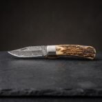 Damascus steel folding pocket knife with stag horn handle and steel bolster, opened and resting on dark slate