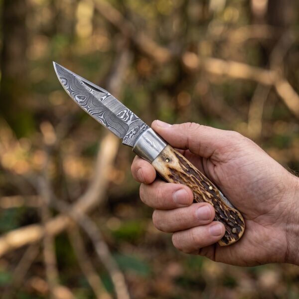 Damascus steel folding pocket knife with stag horn handle held open in a hand against blurred autumn woodland