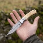 Open Damascus folding knife with natural bone handle resting on an open palm in front of a stone wall and green field