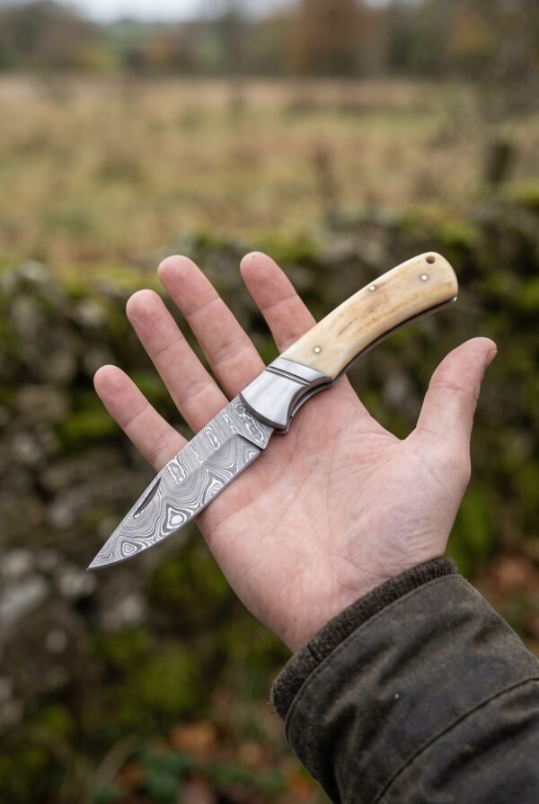 Open Damascus folding knife with natural bone handle resting on an open palm in front of a stone wall and green field