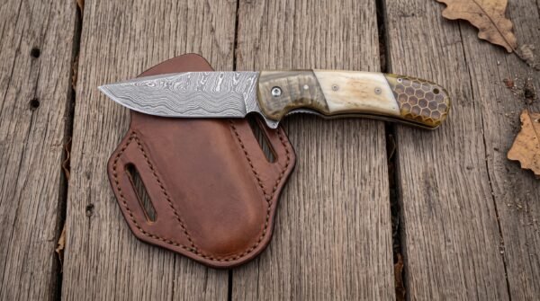 Damascus steel folding pocket knife with ram horn and bone handle resting on its brown leather pancake sheath on planks