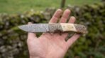 Damascus steel folding knife with ram horn and bone handle held flat in an open hand in front of a mossy stone wall