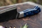 Close up of a high carbon chef knife heel with forged black finish, Damascus bolster and blue resin handle on a dark board