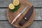Damascus fillet knife with copper handle on a round wooden board with lemon halves, dill and a dark leather sheath