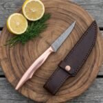 Damascus fillet knife with copper handle on a round wooden board with lemon halves, dill and a dark leather sheath