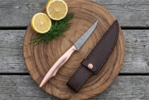 Damascus fillet knife with copper handle on a round wooden board with lemon halves, dill and a dark leather sheath
