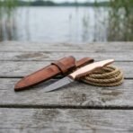 Damascus fillet knife with polished copper handle on a lakeside dock beside a coiled rope and leather sheath
