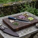 High carbon chef knife with blue resin handle on an end grain board beside chopped onion, celery and its leather sheath